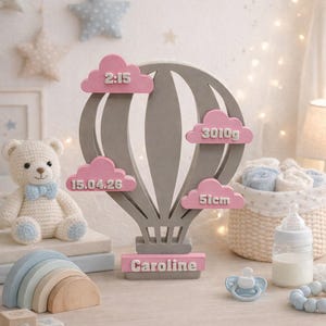 Könnte beinhalten: Ein graues Holz-Andenken in Form eines Heißluftballons mit rosa Wolkenakzenten, das Baby-Statistiken anzeigt: Zeit, Gewicht, Länge und Datum. Der Name "Caroline" ist an der Basis zu sehen. Ein Teddybär, ein Holzregenbogen und Babyzubehör sind ebenfalls vorhanden.