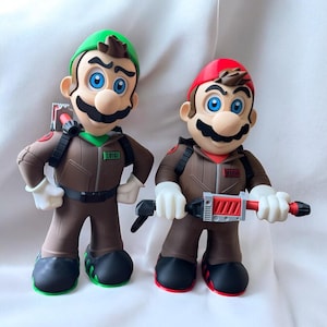 Puede incluir: Dos figuras de personajes de videojuegos, una en verde y otra en rojo, ambas con monos marrones y mochilas de protones. La figura vestida de verde tiene un gorro verde, mientras que la figura vestida de rojo tiene un gorro rojo. Ambas figuras tienen ojos azules y bigote negro.