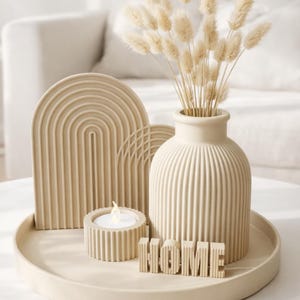 Può includere: Un vassoio decorativo beige contiene un vaso con fiori secchi, un portacandele con una candela accesa e un arco decorativo. La parola "HOME" è scritta in lettere singole. Gli oggetti sono disposti su un tavolo bianco.