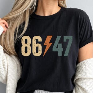 以下が含まれることがあります： 黒のリブ編みクルーネックTシャツ。ベージュの数字「86」、オレンジ色の稲妻、ティール色の数字「47」がプリントされています。グラフィックは、ヴィンテージ風の加工が施されています。