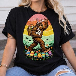 May include: Black t-shirt featuring a Bigfoot character wearing bunny ears and sunglasses, carrying a basket of Easter eggs and a white rabbit. The design includes colorful eggs, flowers, and butterflies, set against a sunset-colored egg shape.