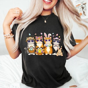 May include: Black t-shirt featuring a graphic of five cats dressed for Easter. The cats wear sunglasses, hats, and bunny ears, holding Easter eggs and baskets. A small bunny with the text "Happy Easter" is also present.
