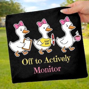 May include: Black tote bag featuring three cartoon ducks with pink bows. One duck holds a clipboard, another a pen, and the third a mug. Text reads "Off to Actively Monitor" in yellow and pink.