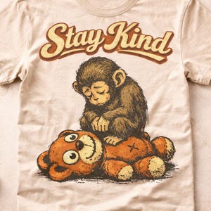 Pode incluir: Camiseta bege com um desenho de um macaco marrom sentado em um urso de pelúcia marrom. O texto "Stay Kind" está arqueado acima do macaco. O design tem um estilo vintage desenhado à mão.