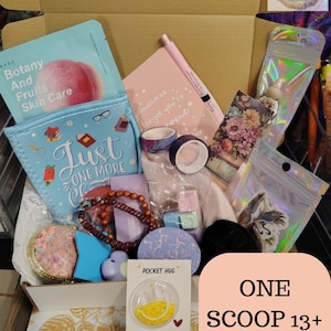 May include: A box filled with various items, including skincare products, a pen, a notebook, washi tape, a bracelet, and a 'Pocket Hug' card. The box has a light brown color and contains a variety of colorful items. Text on the box reads "ONE SCOOP 13+ ITEMS."
