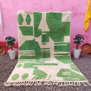 May include: A rectangular area rug featuring a cream-coloured base with bold, abstract geometric patterns in vibrant green. The design includes various shapes, lines, and blocks of colour, finished with a fringe along the bottom edge of the rug.