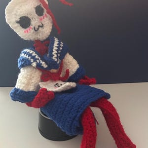 May include: A handmade crochet doll featuring a white skull-shaped head with black eye sockets and a small mouth, wearing a blue and white sailor-style collar over a blue skirt. The doll has long red yarn limbs and a red bow on its head.