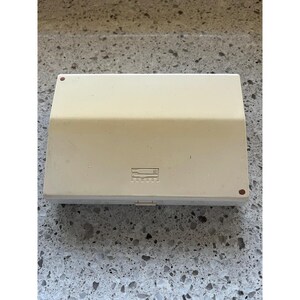 May include: A rectangular, off-white plastic storage case with a hinged lid. The top surface features a small, embossed icon depicting a fork, knife, and spoon. There are two small, reddish-brown circular markings on the top corners of the lid.