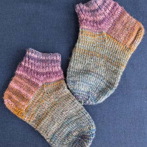 May include: A pair of hand-knitted socks with a colorful, speckled design. The socks feature a mix of purple, pink, orange, and blue hues. The cuff and heel are a darker shade of purple, while the body transitions to a mix of colors.