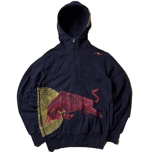 Op de afbeelding: Marineblauwe hoodie met ritssluiting en een rode stierafbeelding op de voorkant. De hoodie heeft een capuchon en een klein Red Bull-logo op de rechterbovenborst. De stierafbeelding is gedetailleerd met rode en gouden accenten.
