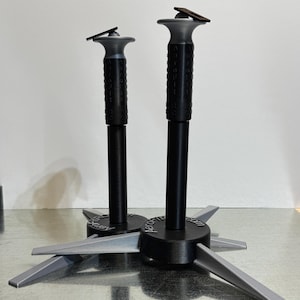 May include: Two black and gray support stands with a star-shaped base. The stands have a cylindrical black pole with a textured grip and a gray top. The base has gray, angled legs. The stands are on a metallic surface.