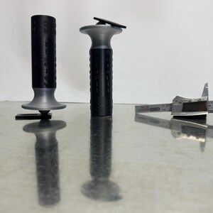 May include: Two black and gray cylindrical objects with text, and a silver caliper are displayed on a reflective surface. The cylindrical objects have a flared base. The caliper is open, showing its measurement scale.
