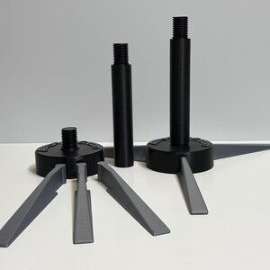 May include: A collection of black and gray leveling feet and shims. The feet have a circular base with a threaded post. Gray shims are arranged around the feet. The items are on a white surface against a gray background.