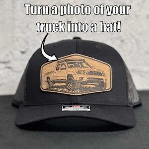May include: A black baseball cap featuring a tan, hexagonal-shaped patch with a laser-engraved illustration of a pickup truck. The text 'Turn a photo of your truck into a hat!' is displayed above the patch, which is centered on the front panel.