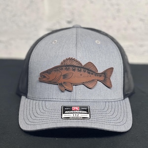 May include: Gray trucker hat with a brown fish patch. The hat has a black mesh back and a curved brim. The patch features a detailed illustration of a fish. The Richardson brand label is visible on the front.