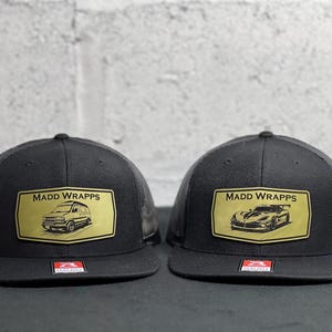 May include: Two black trucker hats with mesh backs and flat brims. Each hat features a rectangular gold patch with the text MADD WRAPPS. The left hat displays a van graphic, while the right hat displays a sports car graphic. Both have a small red logo sticker.