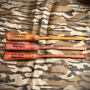 May include: Three wooden duck calls with the text "BURNT TIMBER CUSTOMS" on the barrel. The calls are various shades of brown and red, with a camouflage background. The calls are designed for hunting.