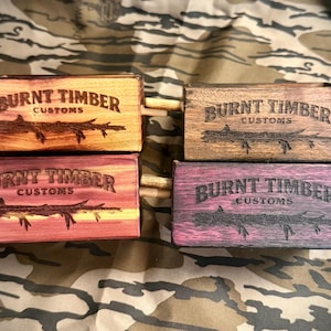 May include: Four wooden boxes with the text "BURNT TIMBER CUSTOMS" and a tree branch design. The boxes are in shades of brown, red, and purple. Each box has a wooden dowel on each side.