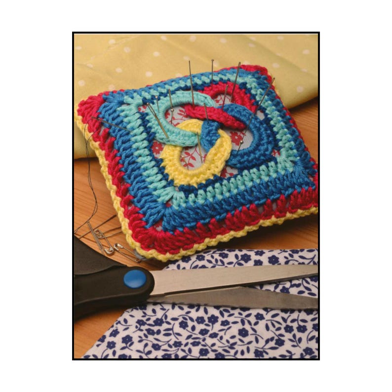 Op de afbeelding: Een vierkant speldenkussen met een kleurrijke gehaakte hoes met concentrische lagen van geel, blauw, turquoise en roze garen. Het midden toont een geknoopt ontwerp en er zitten verschillende metalen spelden in de bovenkant gestoken.