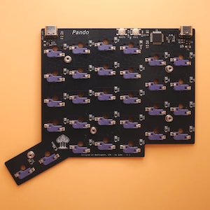 May include: A black printed circuit board for a mechanical keyboard labeled Pando, featuring numerous purple switch sockets, two USB-C ports at the top, and a small tree logo. The board includes various electronic components and surface-mounted parts.