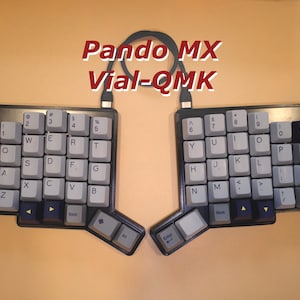 May include: A split ergonomic mechanical keyboard with light gray and dark blue keycaps, featuring the text Pando MX Vial-QMK centered above the two halves. The keyboard is connected by a black cable, with a dark frame and a non-standard layout.