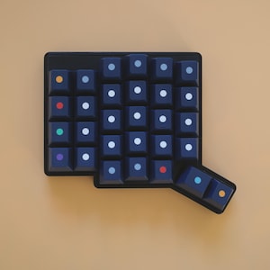May include: A dark navy blue mechanical keyboard macro pad featuring a grid of square keys. Each key is marked with a small, centered circular dot in various colors including white, orange, red, green, purple, and light blue, arranged in a layout.