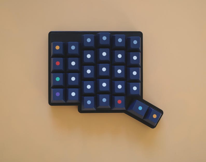 May include: A dark navy blue mechanical keyboard macro pad featuring a grid of square keys. Each key is marked with a small, centered circular dot in various colors including white, orange, red, green, purple, and light blue, arranged in a layout.