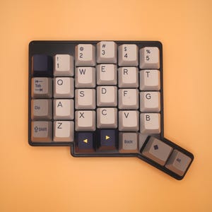May include: A black mechanical keyboard with a non-standard, ergonomic layout. The keycaps are a mix of light gray, dark gray, and black, featuring white alphanumeric characters, symbols, and function labels like Tab, Shift, Back, and Alt.
