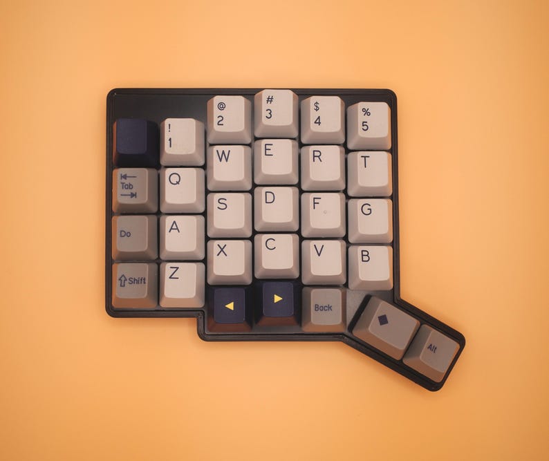 May include: A black mechanical keyboard with a non-standard, ergonomic layout. The keycaps are a mix of light gray, dark gray, and black, featuring white alphanumeric characters, symbols, and function labels like Tab, Shift, Back, and Alt.