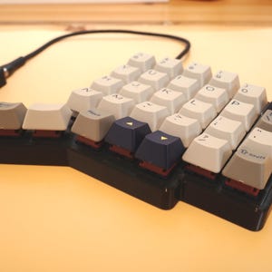 May include: A split ergonomic mechanical keyboard with a black base and a mix of off-white, grey, and dark blue keycaps. The keys feature printed letters, symbols, and the text Help and Shift. A black cable connects the two keyboard halves.