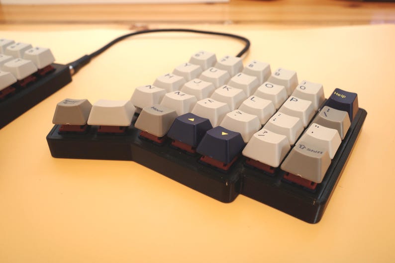 May include: A split ergonomic mechanical keyboard with a black base and a mix of off-white, grey, and dark blue keycaps. The keys feature printed letters, symbols, and the text Help and Shift. A black cable connects the two keyboard halves.