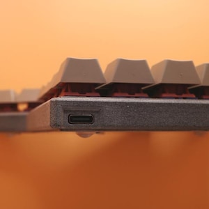 May include: A mechanical keyboard featuring a textured dark gray frame with a visible USB-C port on the side. The keyboard has light gray keycaps with a single orange keycap visible on the right side, mounted on dark red mechanical switches.