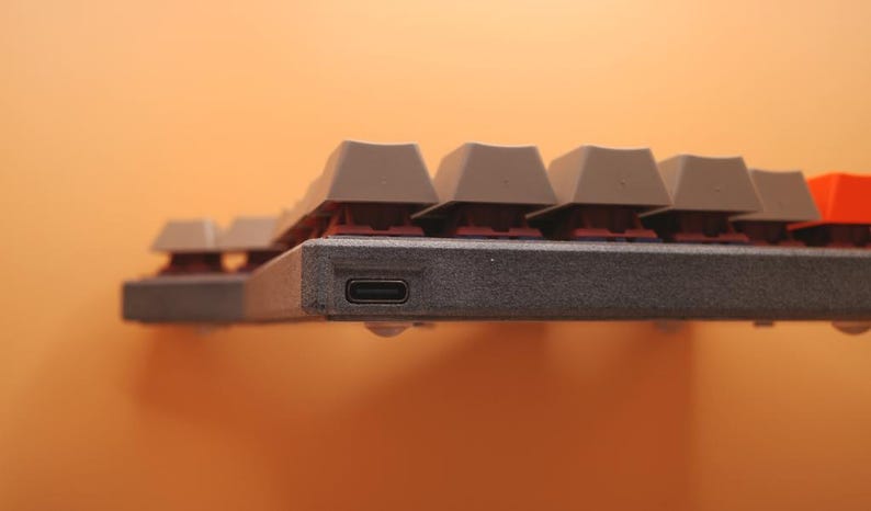 May include: A mechanical keyboard featuring a textured dark gray frame with a visible USB-C port on the side. The keyboard has light gray keycaps with a single orange keycap visible on the right side, mounted on dark red mechanical switches.