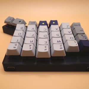 May include: A compact mechanical keyboard with a black base and two USB-C ports. The keycaps are a mix of light gray, dark blue, and white, featuring alphanumeric characters, symbols, and directional arrows printed in black on the top surfaces.