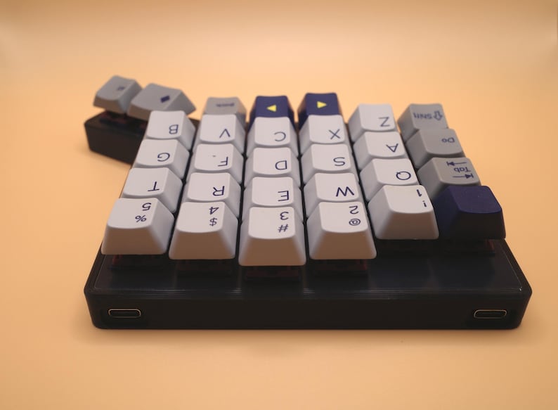 May include: A compact mechanical keyboard with a black base and two USB-C ports. The keycaps are a mix of light gray, dark blue, and white, featuring alphanumeric characters, symbols, and directional arrows printed in black on the top surfaces.
