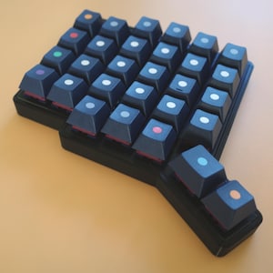 May include: A black ergonomic split mechanical keyboard featuring dark keycaps with small, centered circular icons in various colors including white, orange, red, green, purple, pink, and light blue, arranged in a staggered, non-rectangular layout.