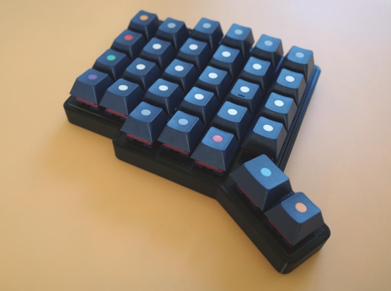May include: A black ergonomic split mechanical keyboard featuring dark keycaps with small, centered circular icons in various colors including white, orange, red, green, purple, pink, and light blue, arranged in a staggered, non-rectangular layout.