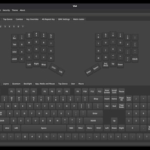 May include: A dark gray software interface titled Vial displaying a split ergonomic keyboard layout on the top half and a standard full-size keyboard layout on the bottom half, with various menu tabs including Keymap, Macros, and QMK Settings.