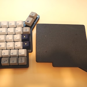 May include: A split mechanical keyboard half featuring white, gray, and dark blue keycaps with QWERTY layout lettering, alongside a separate black rectangular base plate with an extended side arm, both resting on a light surface.