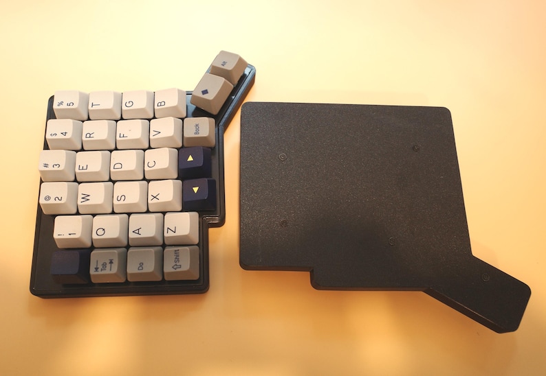 May include: A split mechanical keyboard half featuring white, gray, and dark blue keycaps with QWERTY layout lettering, alongside a separate black rectangular base plate with an extended side arm, both resting on a light surface.