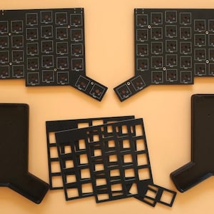 May include: Two black printed circuit boards with square switch cutouts, two black rectangular keyboard cases with angled extensions, and two black grid-patterned switch plates arranged on a flat surface for a split mechanical keyboard build.