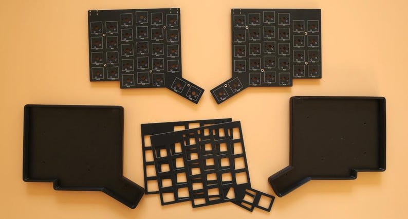 May include: Two black printed circuit boards with square switch cutouts, two black rectangular keyboard cases with angled extensions, and two black grid-patterned switch plates arranged on a flat surface for a split mechanical keyboard build.