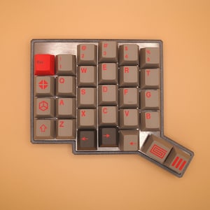 May include: A mechanical keyboard macro pad with a metallic frame, featuring grey keycaps with red legends and a single bright red Esc key. The layout includes alphanumeric keys, symbols, and specialized keys with geometric icons and red arrow symbols.