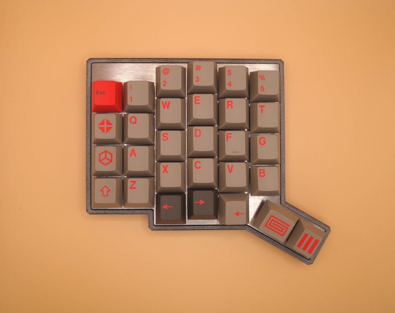 May include: A mechanical keyboard macro pad with a metallic frame, featuring grey keycaps with red legends and a single bright red Esc key. The layout includes alphanumeric keys, symbols, and specialized keys with geometric icons and red arrow symbols.