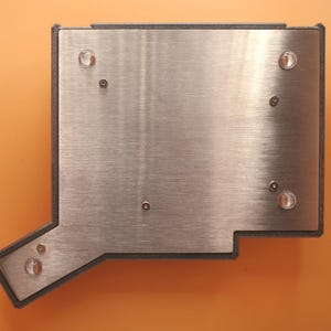 May include: A rectangular metal plate with an irregular extension on the bottom left corner, featuring a brushed silver finish. The plate is secured within a dark gray frame and includes four clear circular feet and several small metal screws.