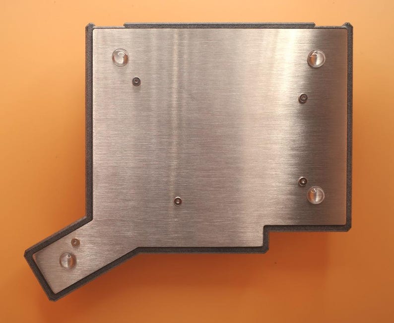 May include: A rectangular metal plate with an irregular extension on the bottom left corner, featuring a brushed silver finish. The plate is secured within a dark gray frame and includes four clear circular feet and several small metal screws.