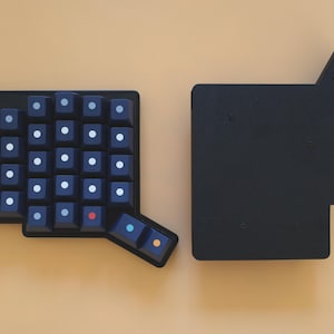 May include: A black split ergonomic mechanical keyboard half featuring a grid of dark keys with white circular legends, alongside a solid black rectangular base plate with an extended corner section, both arranged on a plain light tan surface.