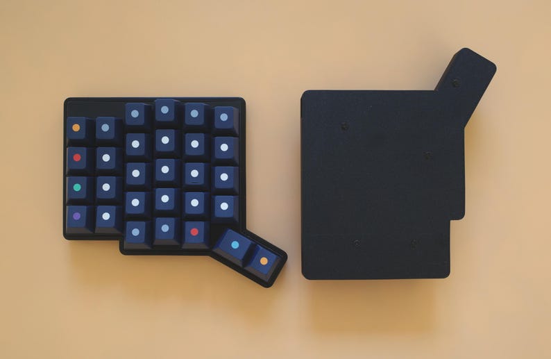 May include: A black split ergonomic mechanical keyboard half featuring a grid of dark keys with white circular legends, alongside a solid black rectangular base plate with an extended corner section, both arranged on a plain light tan surface.