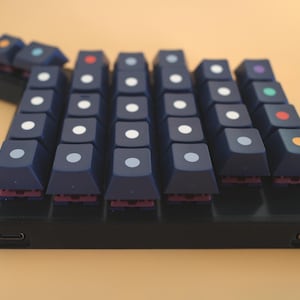 May include: A custom mechanical macro keypad featuring dark blue keycaps with centered circular dots in various colors including white, red, orange, green, and purple. The device has a black rectangular base with two USB-C ports on the front edge.