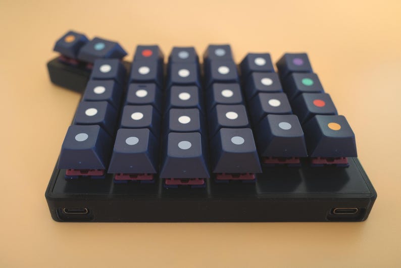 May include: A custom mechanical macro keypad featuring dark blue keycaps with centered circular dots in various colors including white, red, orange, green, and purple. The device has a black rectangular base with two USB-C ports on the front edge.